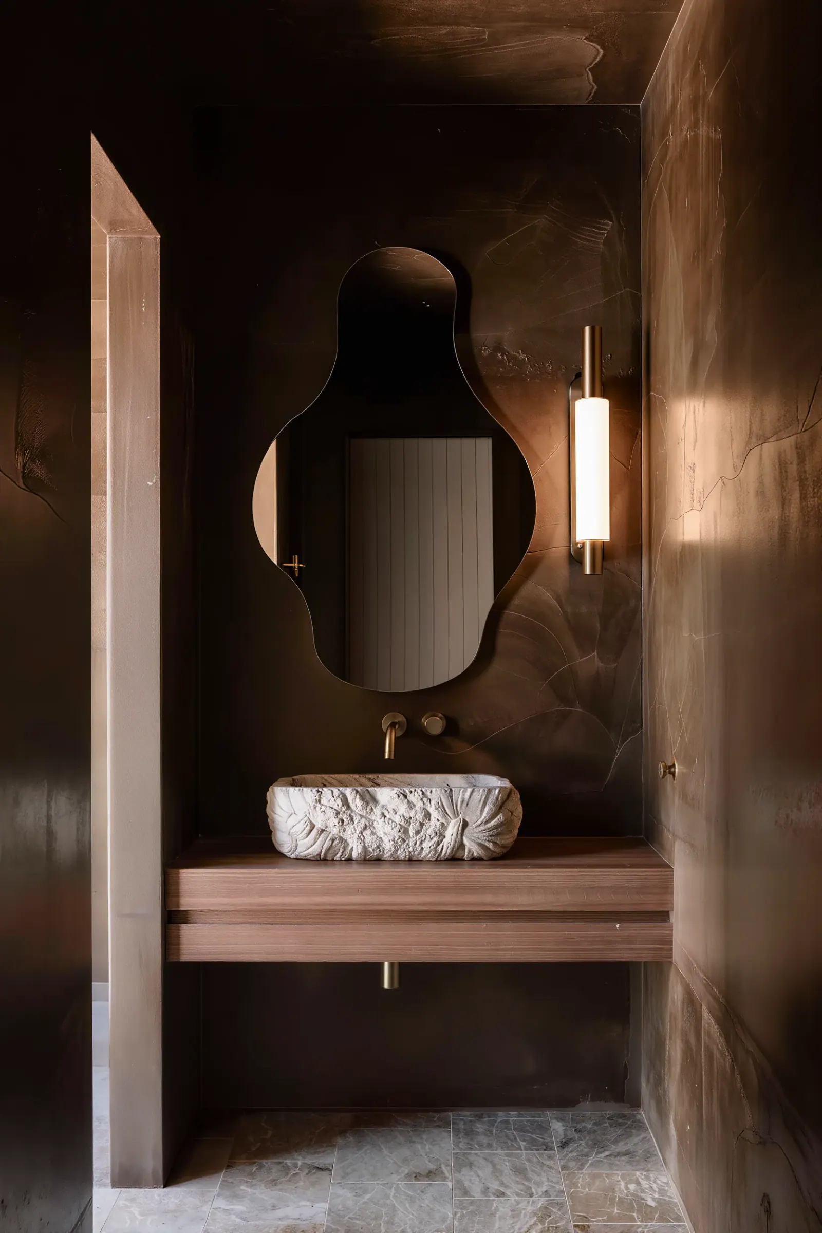 Moody powder bath with dark plaster walls, a carved travertine vessel sink, and a brass sconce