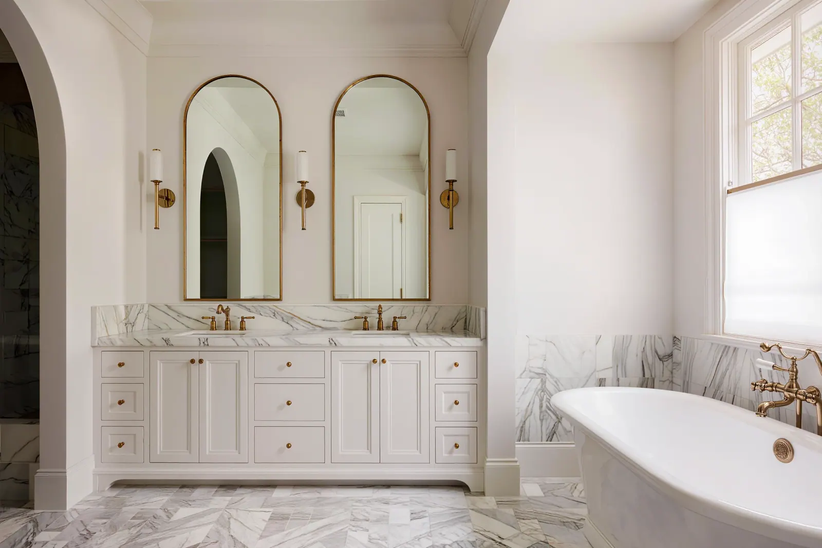 Bright primary bath with a white oak double vanity, arched brass mirrors, and herringbone marble floor